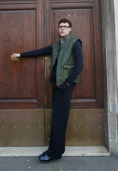 Chaleco acolchado verde sobre una camiseta de manga larga negra, combinado con pantalones negros y zapatos negros brillantes. Apoyado contra una puerta de madera.