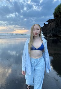 Osoba stojąca na mokrym piasku, ubrana w niebieski bikini, jasnoniebieskie luźne spodnie i otwartą białą koszulę, z warkoczem we włosach, na tle pochmurnego nieba.