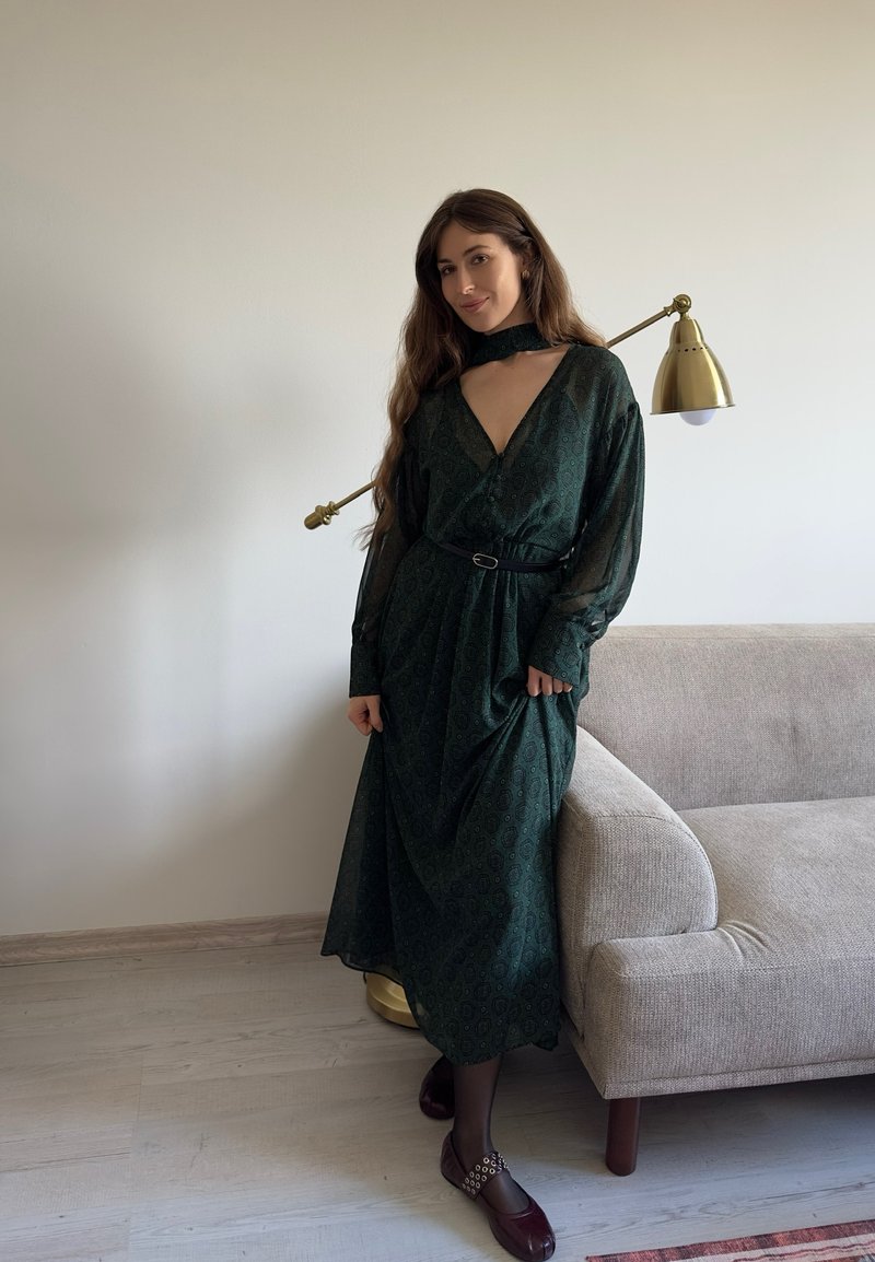 Grøn mønsterkjole med lange ærmer, V-udskæring og et bælte. Sammen med mørk muave sko og en choker. Beige sofa i baggrunden.