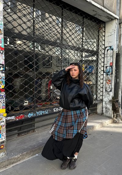 Joven mujer con chaqueta negra y falda de cuadros está en la acera frente a una tienda cerrada con una rejilla de seguridad metálica.
