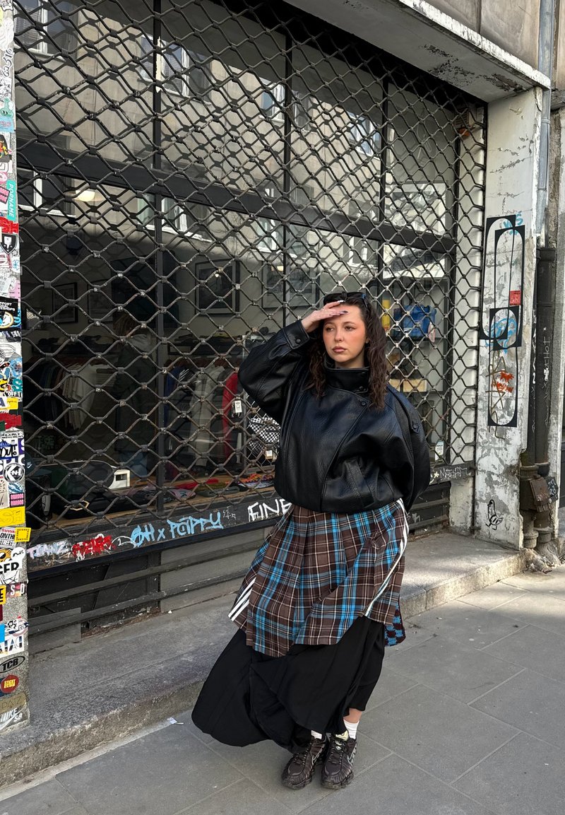 Jeune femme en veste noire et jupe à carreaux se tient sur le trottoir devant un magasin fermé avec une grille de sécurité métallique.