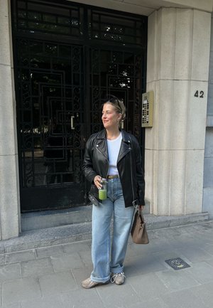 Chaqueta de cuero negra, camisa blanca, vaqueros de mezclilla sueltos de color azul claro, bolso beige, zapatos grises, sosteniendo una bebida verde, de pie junto a una pared de edificio texturada.