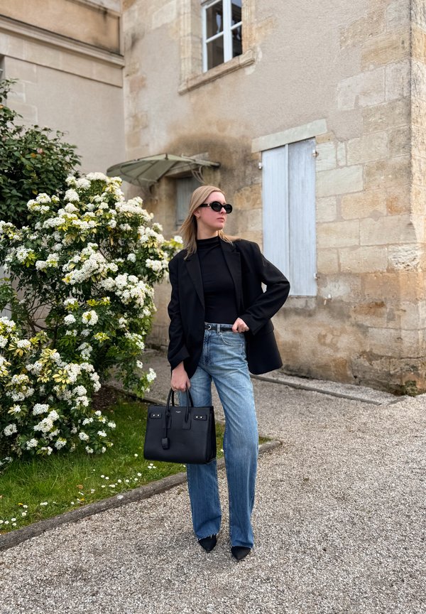 Kvinde iført sort blazer og solbriller, blå jeans, holder en sort håndtaske, står nær en blomstrende busk og en stenhøj bygning med skodderne lukkede.