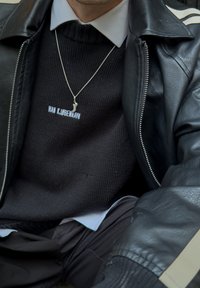 Sweater de malha preto com o texto "HAN KJØBENHAVN", sobreposto a uma jaqueta de couro preta com fecho de zíper, e um colar prateado com um pequeno pingente.