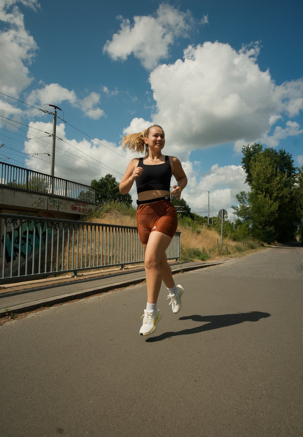 Juodos sportinės marškinėliai ir oranžinės šortai, su baltomis sportinėmis bateliais. Bėga asfaltuotu taku po mėlynu dangumi su debesimis ir šalia augmenijos.