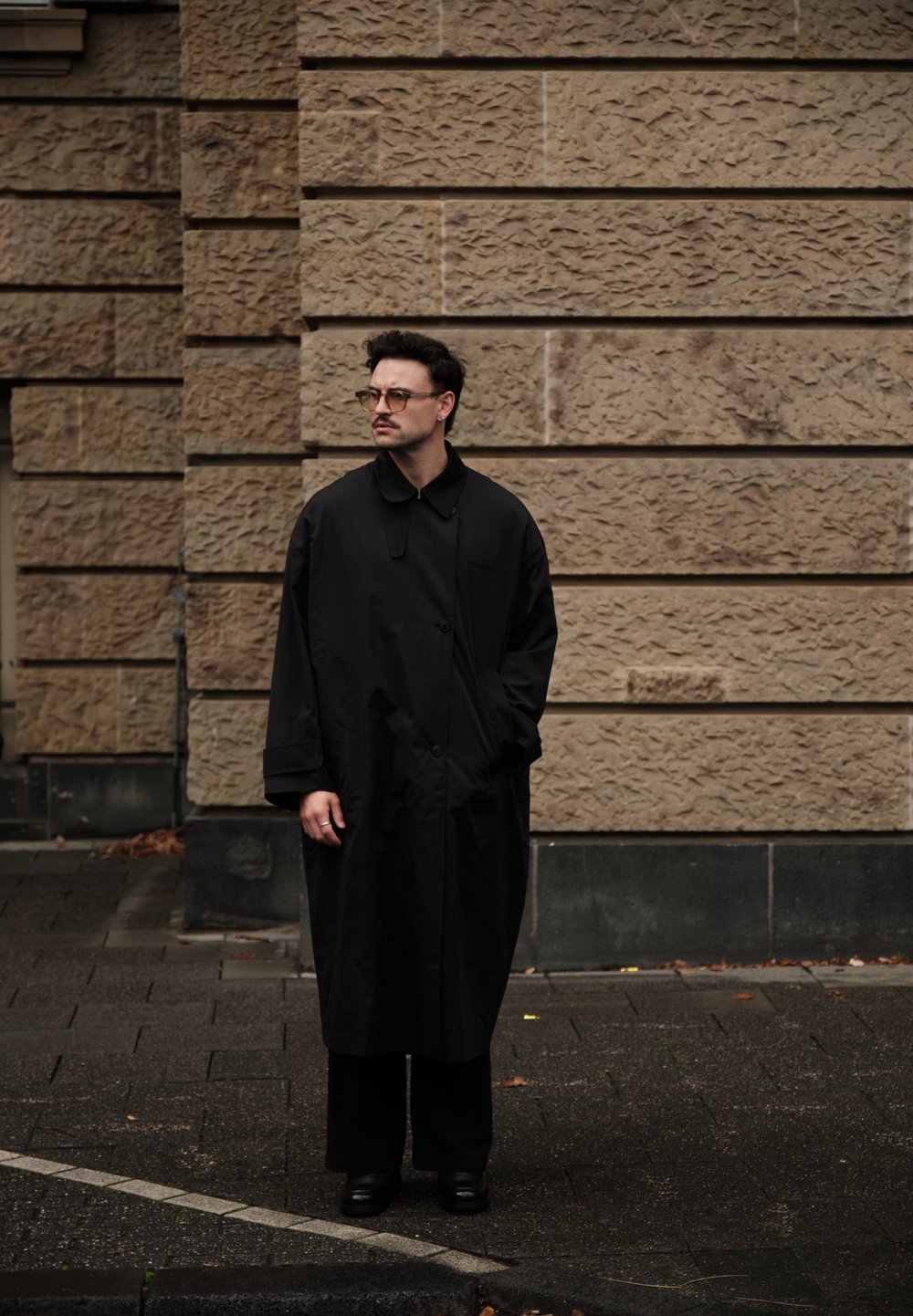 Mustast keskmise pikkusega mantel kõrge kraega, kinnitatav ühe nööbiga, kantud mustade pükste ja kingade peal. Taustal tekstuuriga kivimüür.