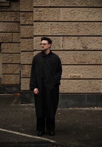 Mustast keskmise pikkusega mantel kõrge kraega, kinnitatav ühe nööbiga, kantud mustade pükste ja kingade peal. Taustal tekstuuriga kivimüür.