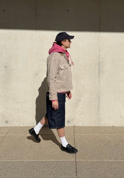 Hombre caminando junto a una pared de hormigón vestido con una sudadera con capucha rosa, chaqueta beige, pantalones cortos oscuros, calcetines blancos, zapatos negros, gorra y gafas de sol.