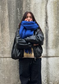 Čierna kožená bomberová bunda, nadrozmerný modrý �šál, tmavé džínsy a textúrovaná čierna kabelka na pozadí šedej steny.