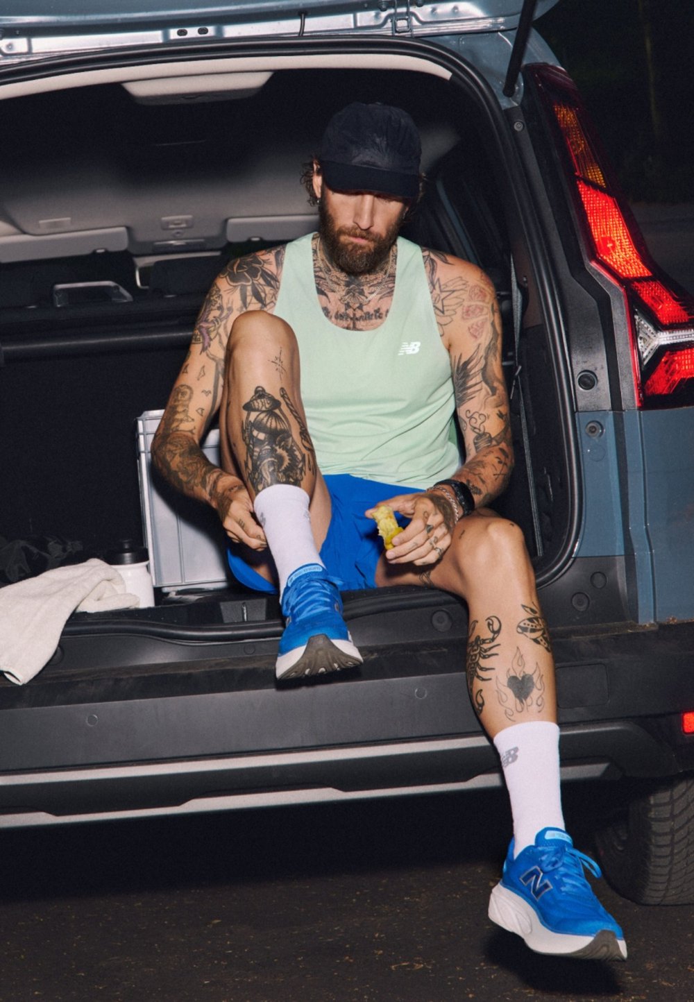 Mėlyni sportiniai bateliai, šviesiai žalias marškinėliai be rankovių ir mėlynos šortai. Tatuiruotės ant rankų ir kojų. Sėdi atidarytame automobilio bagažinė.
