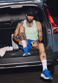 Mėlyni sportiniai bateliai, šviesiai žalias marškinėliai be rankovių ir mėlynos šortai. Tatuiruotės ant rankų ir kojų. Sėdi atidarytame automobilio bagažinė.