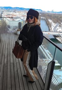 Femme portant un manteau noir, un chapeau en fourrure, des lunettes de soleil et un pantalon Adidas marron, se tenant sur un balcon surplombant une ville enneigée et une rivière.