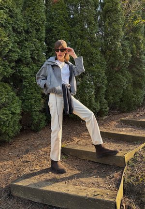 Mujer con gafas de sol, chaqueta a cuadros, pantalones blancos y botas marrones, de pie al aire libre sobre unos escalones de madera junto a altos árboles perennes.
