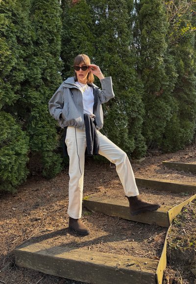 Mujer con gafas de sol, chaqueta a cuadros, pantalones blancos y botas marrones, de pie al aire libre sobre unos escalones de madera junto a altos árboles perennes.