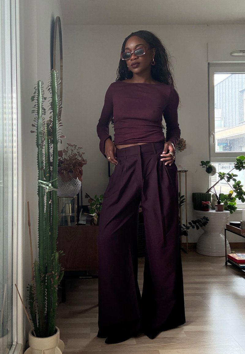 Bordeauxrotes Langarmoberteil und weit geschnittene Hosen aus glattem Stoff, mit tailliertem Schnitt und fließendem Design; ergänzt durch Sonnenbrille und Accessoires.