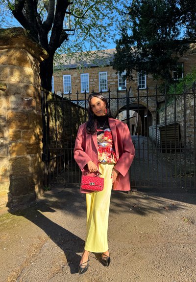 Mujer de pie al aire libre cerca de una pared de piedra y una puerta de hierro, vistiendo un abrigo rosa, blusa floral, falda amarilla, zapatos negros y sosteniendo un bolso rojo.