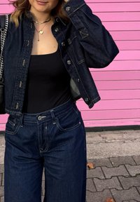 Temno modra jeans jakna z vidnimi šivi, velikimi gumbi in kratkim krojem, združena s črnim oprijetim topom in temnimi visokim pasom jeans hlačami.