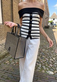 Μαυρόασπρη ριγέ μπλούζα off-the-shoulder με κουμπιά, συνδυασμένη με λευκά παντελόνια με φαρδιά γραμμή και μια πλεκτή τσάντα μαύρη-καφέ.