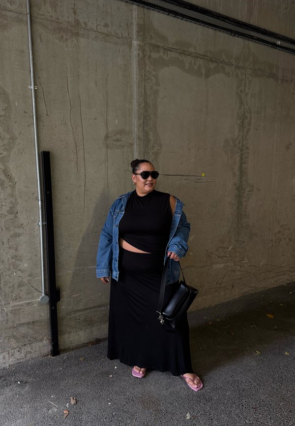 Femme portant une robe noire à découpes, une veste en jean oversized, des lunettes de soleil noires et des sandales roses, tenant un sac à main noir, debout contre un mur en béton.
