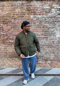 Veste matelassée vert olive avec deux poches avant, portée sur une chemise blanche, associée à un jean ample bleu clair et des baskets grises.