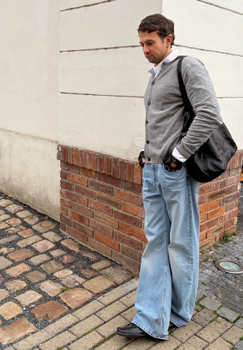 Moški s črno ramensko torbico, sivim jopičem, belo srajco, rokavicami brez prstov, širokimi modrimi kavbojkami in črnimi čevlji stoji na tlakovanem ulici.