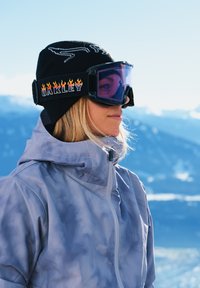 Lunettes de ski avec verres teintés violets, sangle noire avec l'inscription "OAKLEY" et accents de flamme, accompagnées d'une veste tie-dye gris clair et d'un bonnet noir.