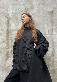 Svart, teksturert, oversized genser med glidelås i halsen, med preget bølgemønster. Kombinert med m�ørke bukser foran en nøytral vegg.