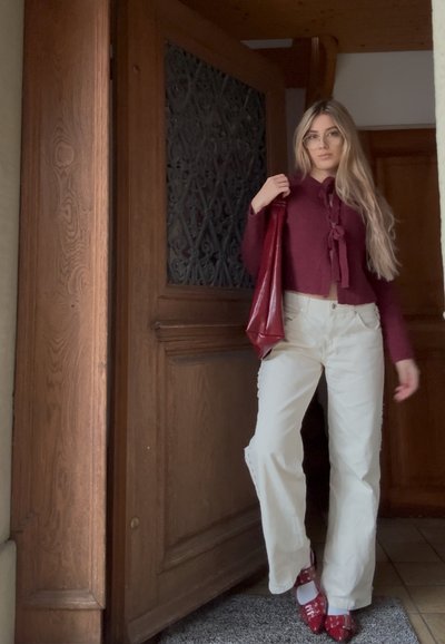 Jersey corto de punto color granate con detalle de lazo, pantalones de pierna ancha color crema y zapatos de tacón rojos con diseño perforado. Fondo de puerta de madera.