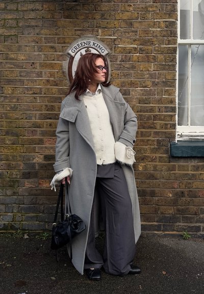 Mujer con abrigo gris y pantalones anchos de pie contra una pared de ladrillos con un cartel de Greene King, sosteniendo un bolso negro y mirando hacia la derecha.