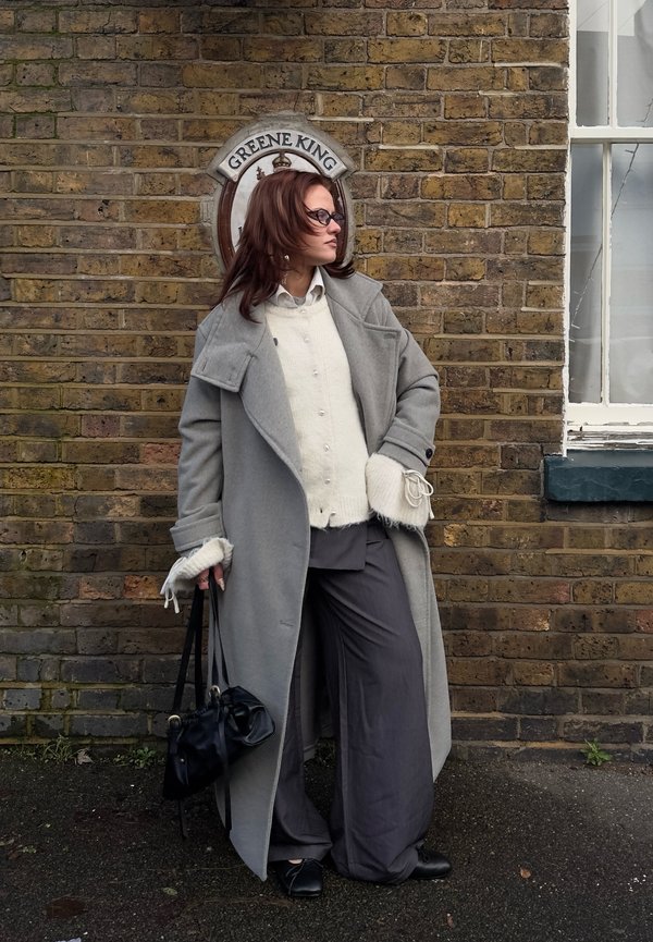 Kvinde iført grå frakke og vide bukser står op ad en murstensvæg med et Greene King-skilt, holder en sort håndtaske og kigger mod højre.