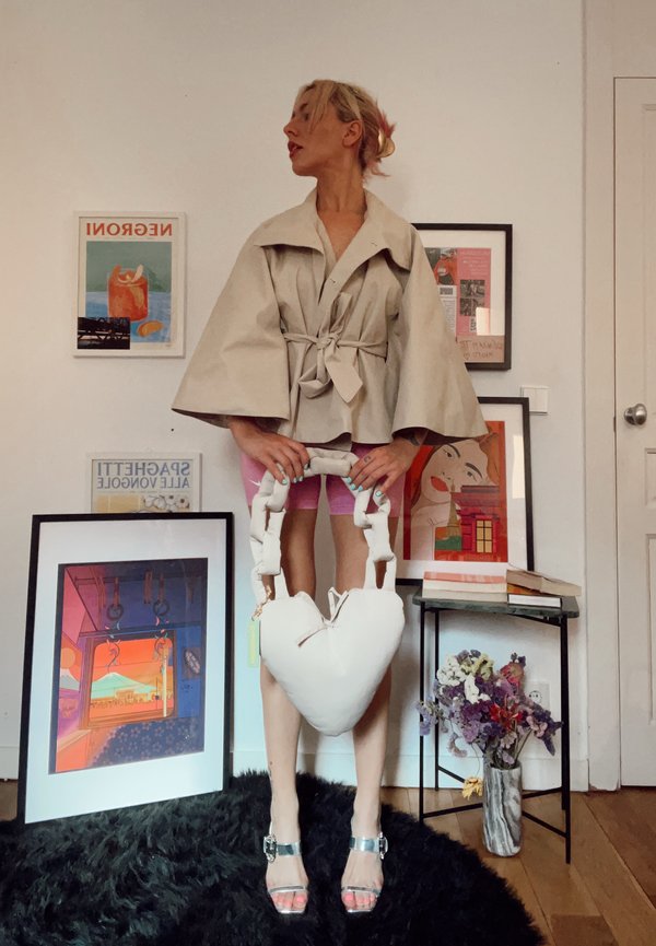 Kvinde i beige jakke og lyserøde shorts holder en stor hvid hjerteformet taske, stående i et rum med indrammet kunstværk og blomster på gulvet.