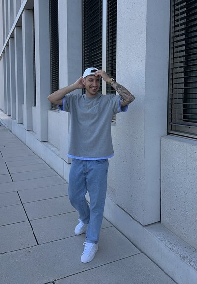 Camisa gris de manga corta con acentos azules, jeans de mezclilla claros y zapatillas blancas. El fondo presenta paredes texturizadas de color claro y ventanas.