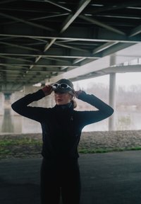 Jong persoon die een honkbalpet met zonnebril erop aanpast onder een brug bij een rivier, gekleed in donkere sportkleding.