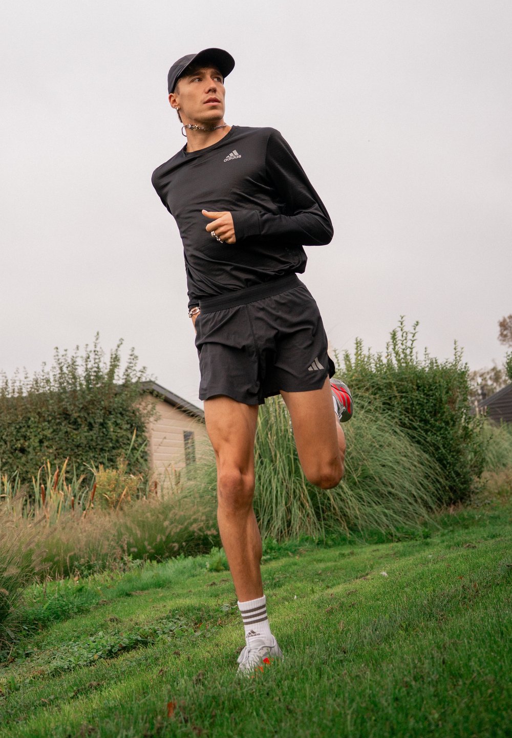 Giovane che corre all'aperto sull'erba, indossando un completo sportivo nero, un cappellino e calze Adidas bianche, con cespugli e piccole case sullo sfondo.