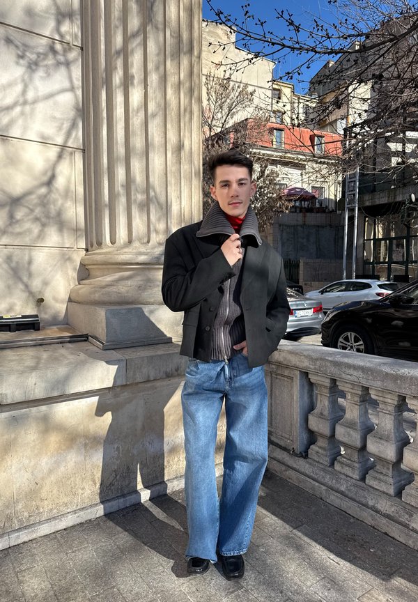 Jonge man staand naast een stenen kolom en balustrade, draagt een zwarte jas, een grijze trui, een rode coltrui, blauwe jeans en zwarte schoenen in een stedelijke omgeving.