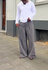 Calças cinzas longas e largas com pregas, combinadas com uma camisa de manga longa tipo henley branca. O calçado inclui sapatos escuros sem atacadores com um padrão.