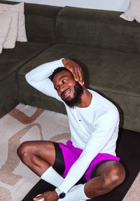 Homme en chemise blanche à manches longues et short violet assis sur un tapis de sol, étirant le cou avec la main sur le côté de la tête près d'un canapé vert.