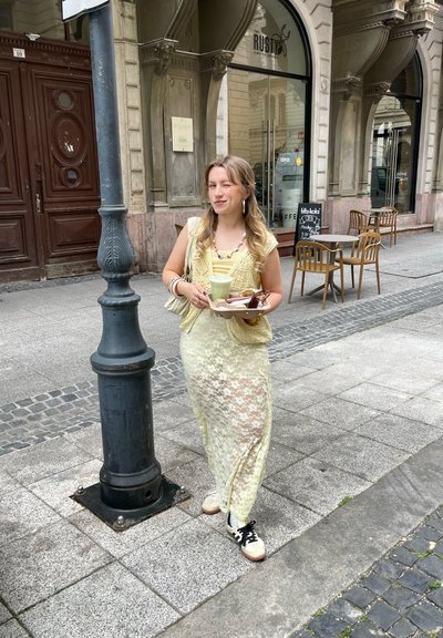 Joven mujer con un vestido de encaje amarillo en capas, sosteniendo una bandeja con bebidas y pasteles. Lleva un chaleco de ganchillo y zapatillas. Calle adoquinada.