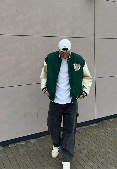 Chaqueta universitaria verde y crema con un logotipo, camiseta blanca, vaqueros oscuros y holgados, y zapatillas blancas, de pie sobre una superficie pavimentada.