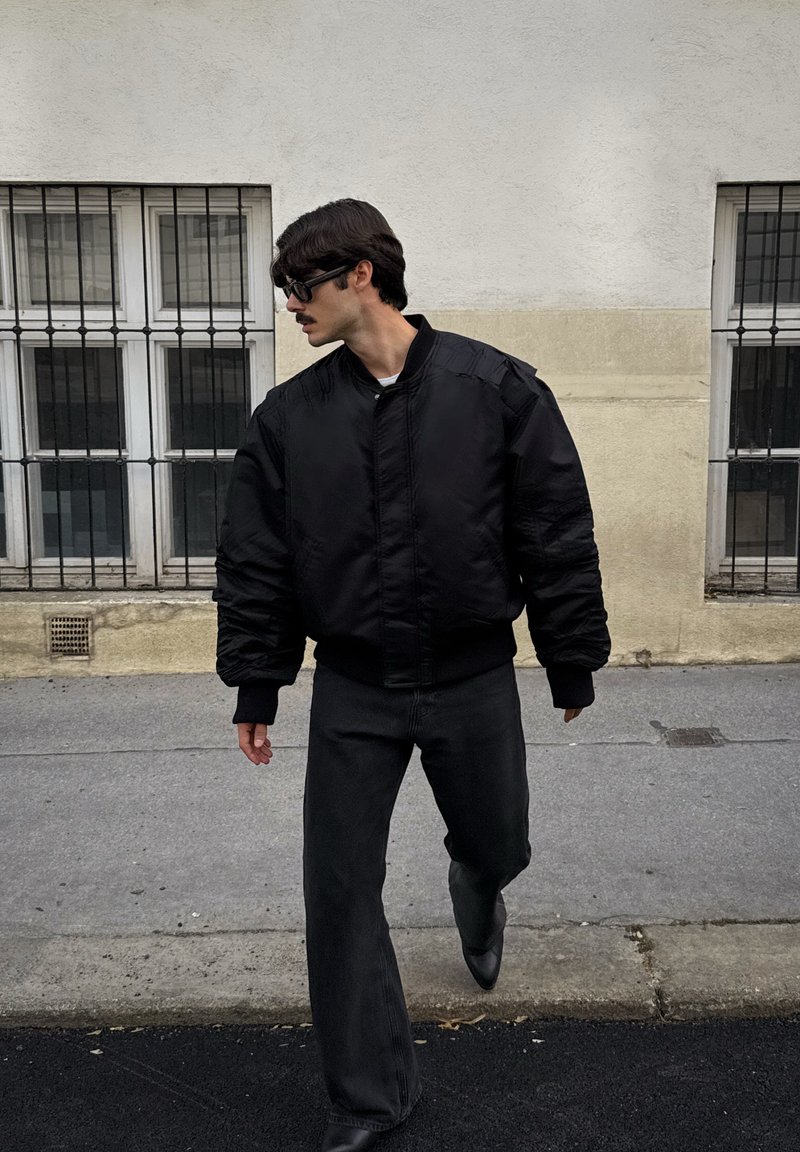 Casaco bomber preto com ombros acolchoados, punhos e cintura canelados, usado sobre uma camisa branca, combinado com calças largas pretas.