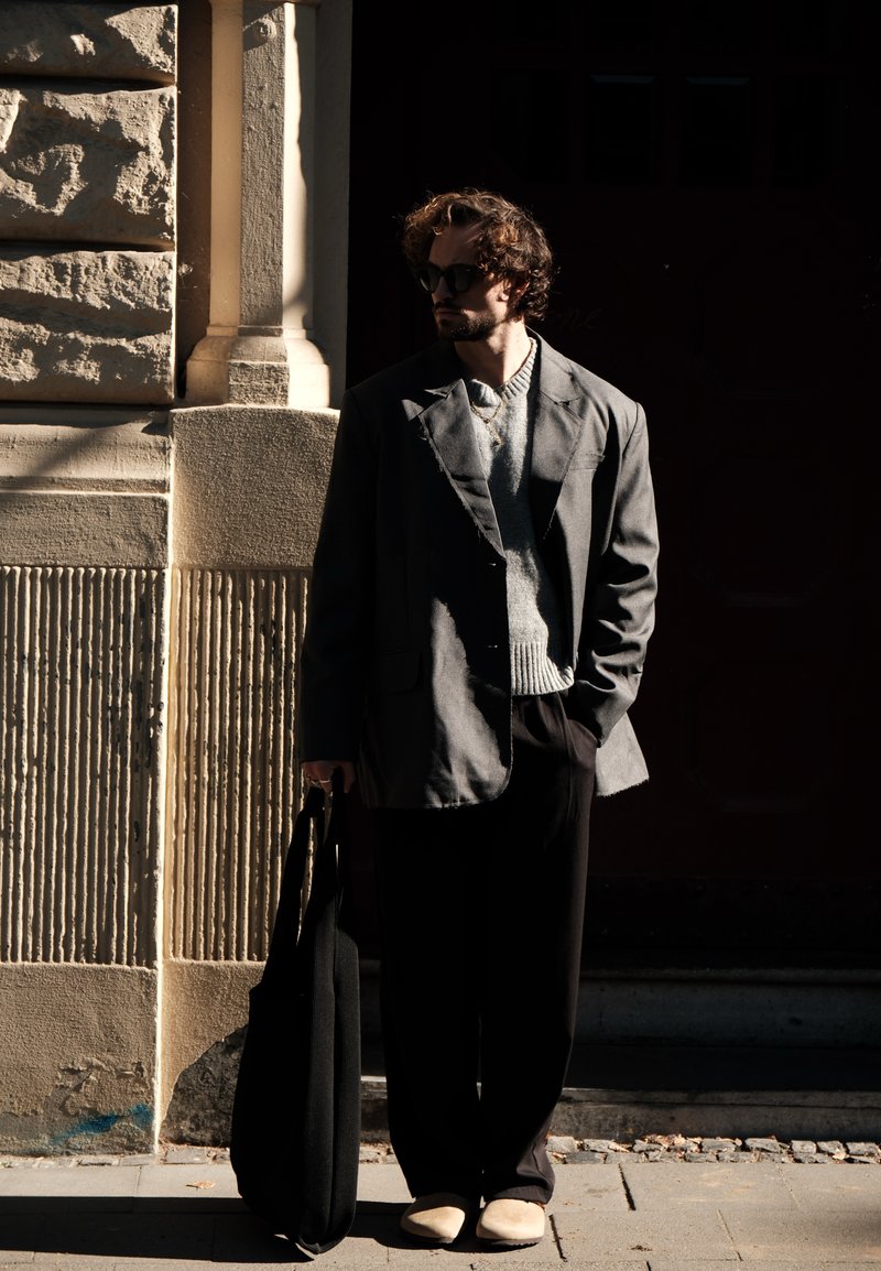 Γκρι oversized σακάκι πάνω από ένα ανοιχτό γκρι πουλόβερ, μαύρο παντελόνι και μπεζ slip-on παπούτσια. Κρατάει μια μαύρη τσάντα tote. Σκιασμένο αστικό φόντο.