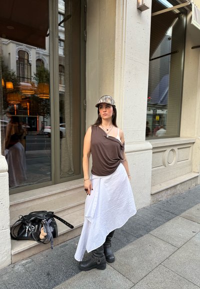 Top asimétrico marrón combinado con una falda blanca fluida. Botines negros con tachuelas. Gorra de camuflaje. Bolsa negra con accesorios en el suelo.