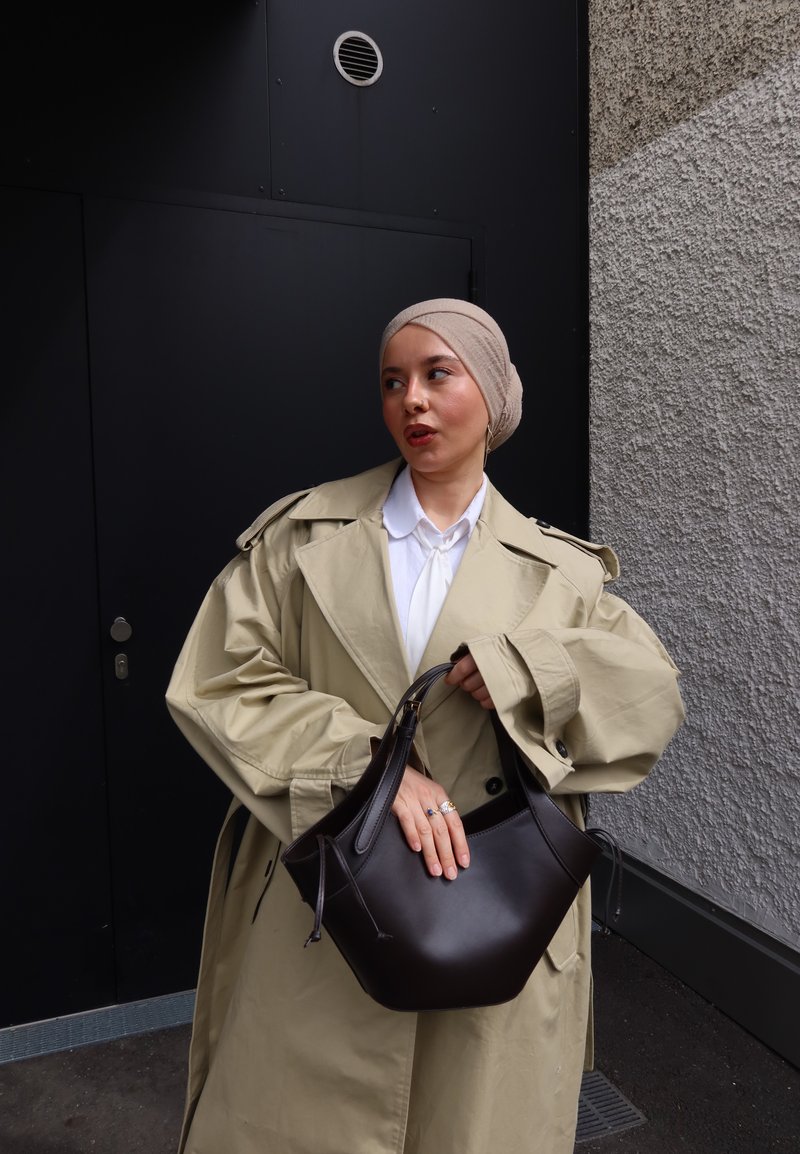 Vrouw die een beige trenchcoat en lichtbruine hoofddoek draagt, houdt een donkerbruine handtas vast en staat tegen een zwarte en gestructureerde grijze muur.