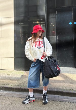 Sudadera blanca con el logo rojo de Nike, culottes de mezclilla azules, sombrero de cubo rojo y zapatillas negras con acentos plateados. Bolsa negra con el texto "AIR".