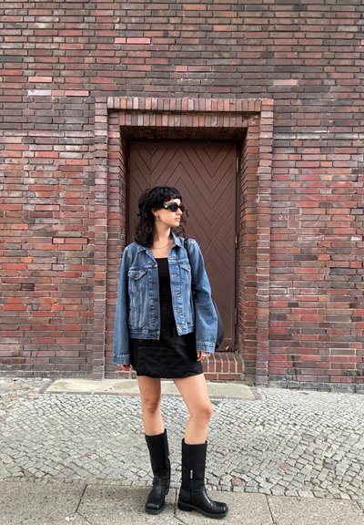 Chaqueta de mezclilla con lavado claro, vestido negro y botas negras hasta la rodilla. El fondo presenta una pared de ladrillo texturizada y una puerta marrón.