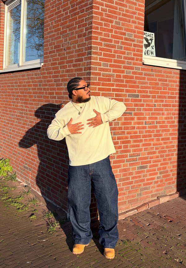 Man met gevlochten haar en bril staat tegen een bakstenen muur, draagt een crèmekleurige trui, wijde spijkerbroek en beige laarzen in het zonlicht.