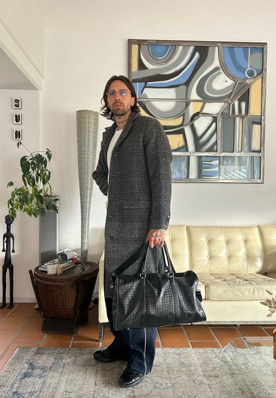 Hombre con cabello a la altura de los hombros, usando gafas y un abrigo gris de cuadros, sosteniendo un bolso negro de cuero trenzado en un ambiente de sala de estar.
