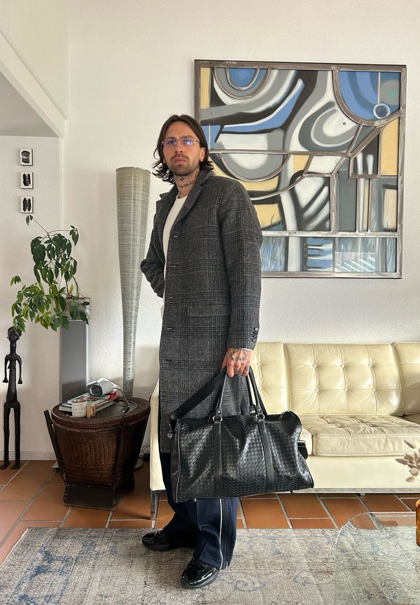 Bărbat cu părul până la umeri, purtând ochelari și o haină gri cu carouri, ținând o geantă neagră din piele țesută, într-un decor de sufragerie.