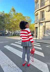 Röd och vit randig tröja, grå plisserad kjol, grå strumpbyxor, röda ballerinat och röd väska. Stadsmiljö med träd och blå himmel.
