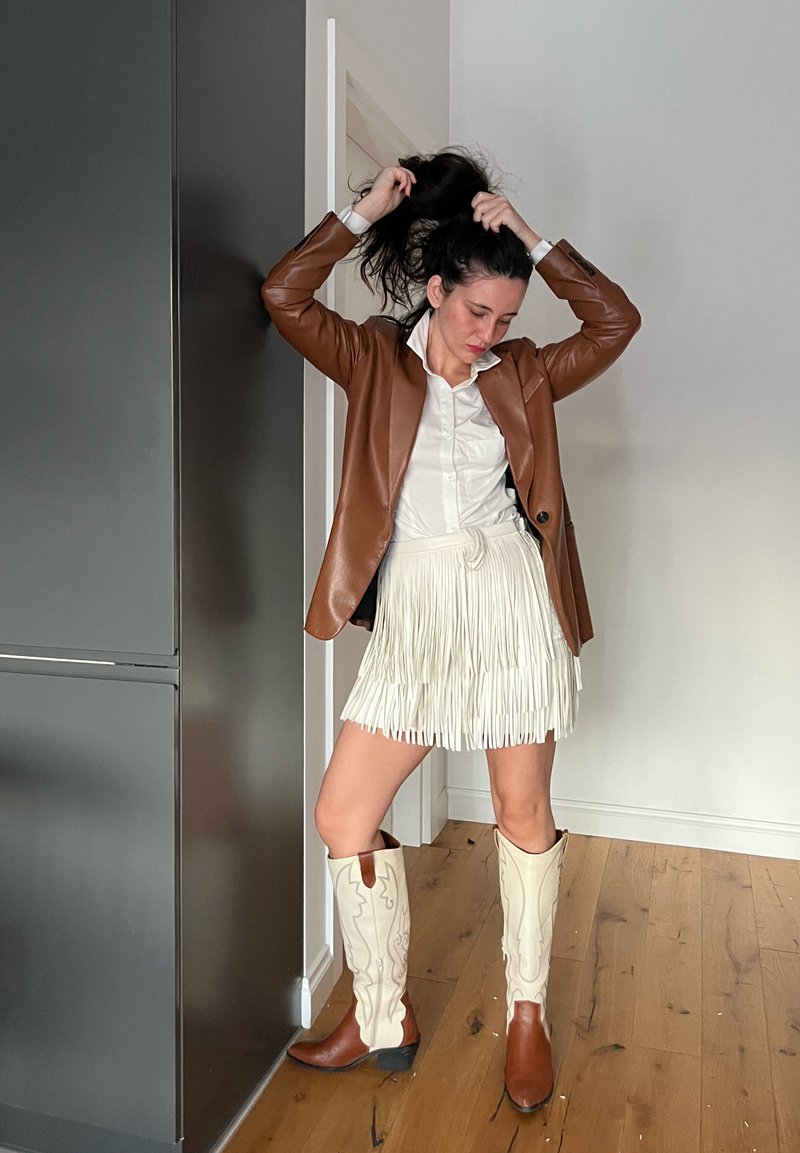 Brūns ādas žakete, balta ar pogām aizdarāma krekla krekls, balta mini svārku ar pušķiem un krēmkrāsas kovboju zābaki ar brūniem akcentiem, uz gaiša fona.